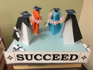 origami penguins stand with graduation caps near a sign reading "Succeed."