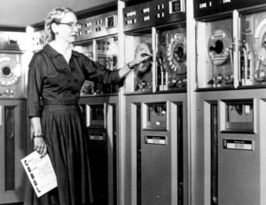Grace Hopper holding a COBOL manual and standing by a Univac II tape drive.