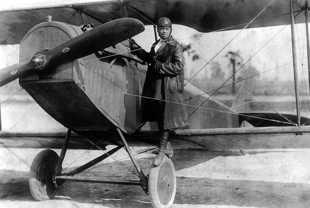 Bessie Coleman stands on one of the tires while holding on to her Curtiss biplane.