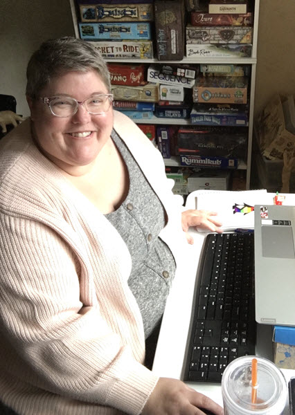 Beth Caldwell smiles at you from her home office. An impressive collection of board games fills a bookshelf next to her desk.