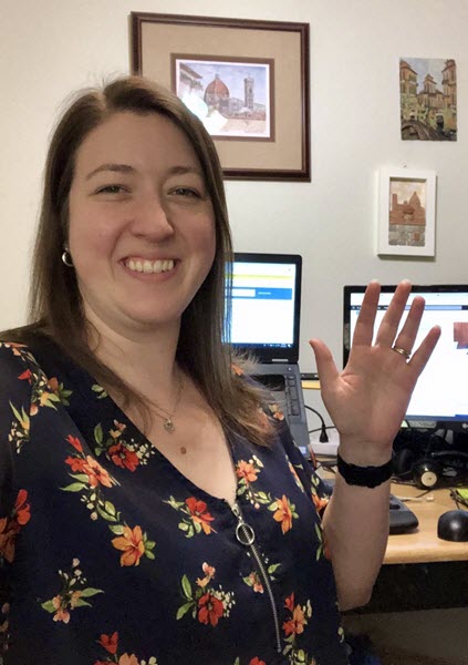 Cheryl Davenport smiles and waves to you from in front of her home office. There is art on the wall behind her and a laptop with an extra monitor.