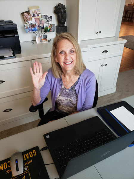 Connie Anderson smiles and waves from her home desk. A black laptop sits on the desk and an arrangement of photos is on a cabinet behind her along with a small sculpture.