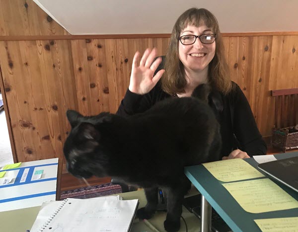 a blurry, black floof of a cat steps on Julie's desk, making herself at home on the top of the desk. Julie waves in the background