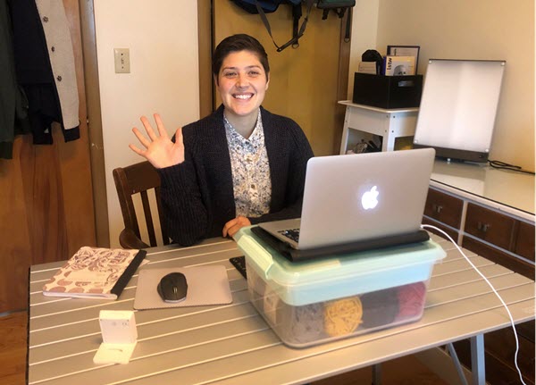 Nicole Langley smiles and waves from her home desk. Her Apple laptop sits atop a Tupperware bin filled with yarn.