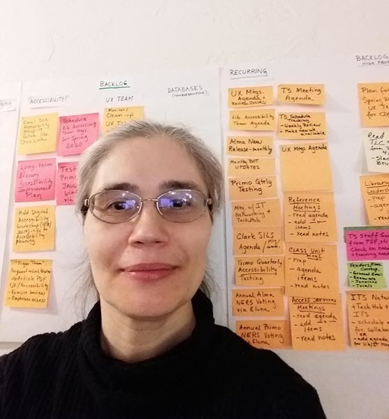 Radka Ballada smiles at you standing in front of her kanban board, which is covered in orange, pink, and green sticky notes.