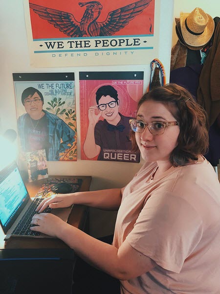 Tasha Roberston smiles at you while seated at her laptop station. Posters next to her feature Lydia X.Z. Brown and Lindsay Amer.