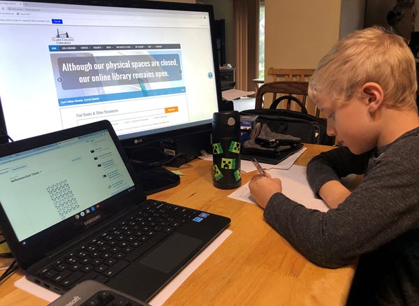 A boy sits at a desk writing on a piece of paper. In front of him on the desk are a laptop, a monitor open to the library homepage, and a Minecraft thermos.