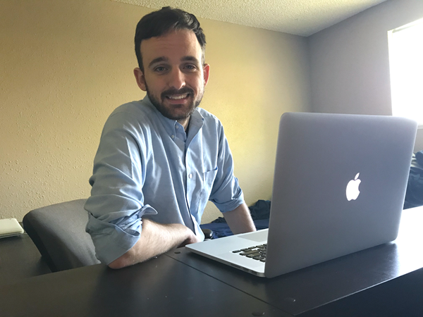 Jonathan Ulrich smiles at you from his home workstation. An apple laptop sits atop his black desk.