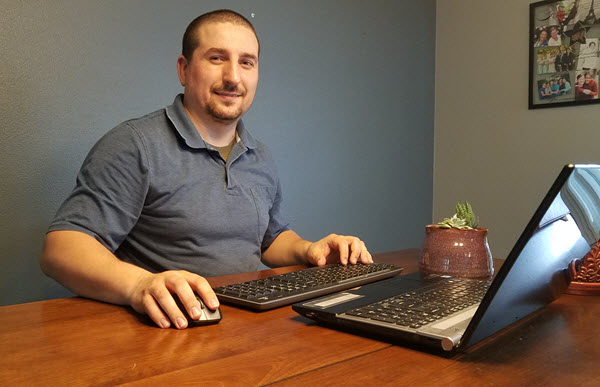 Sergey Kachenkov smiles while sitting at his home desk. He has a wireless mouse and keyboard to go with his laptop, and two succulents keep him company atop his desk.
