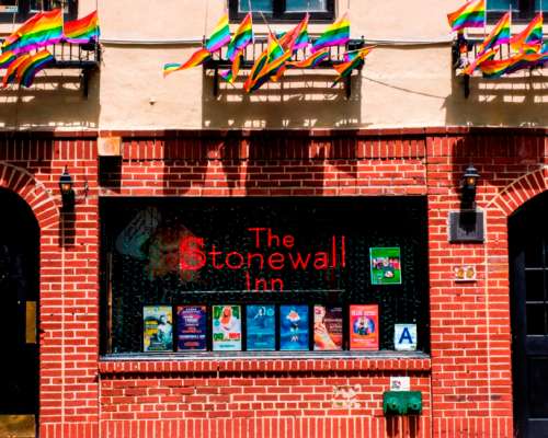 An image of The Stonewall Inn. The building is brick and the name of the inn is in the window. There are rainbow flags above the window.