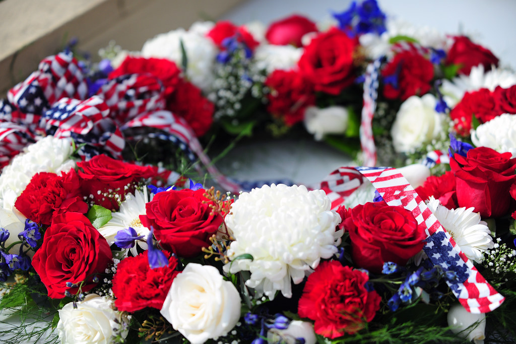 a wreath made from red, white, and blue flowers, with an American flag ribbon