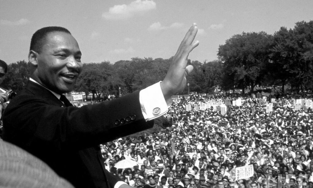 A black and white image of MLK adressing a croud. He is on the left side of the image facing the camera, waving out to the crowd around him