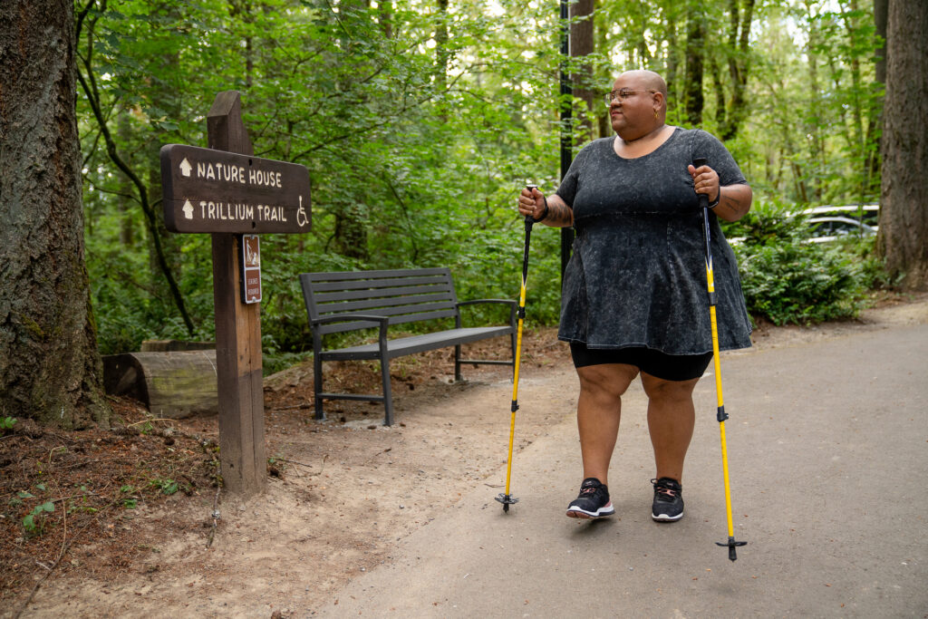 A disabled Black non-binary hiker uses trekking poles and looks at a wooden sign with “Nature House” and “Trillium Trail” marked ahead. The shot is framed with the hiker walking toward the camera and the hiker sports a shaved head, glasses, a gray peplum shirt, black shorts, and black tennis shoes.