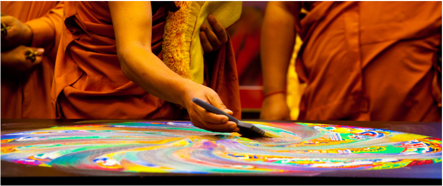 Monks wearing orange sweep away a colorful sand mandala