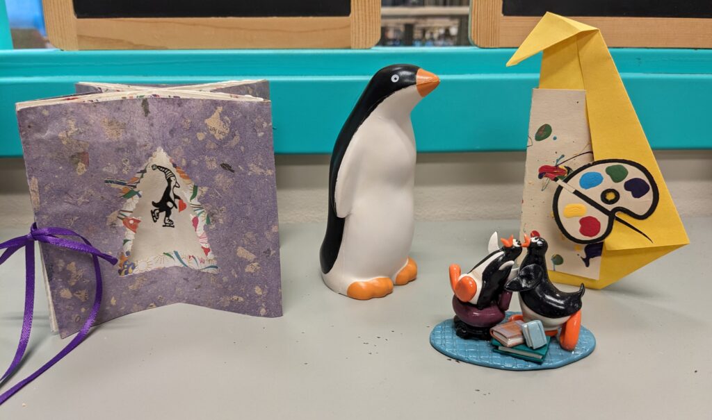 A purple paper card with a hand-drawn penguin, a penguin-shaped stress ball, two clay penguins with books, and a yellow origami penguin with a painter's palette