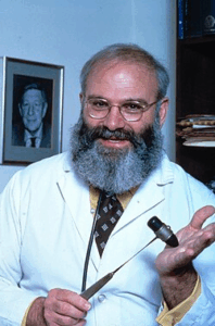 A grey-haired Oliver Sacks with glasses and a beard holding a reflex hammer
